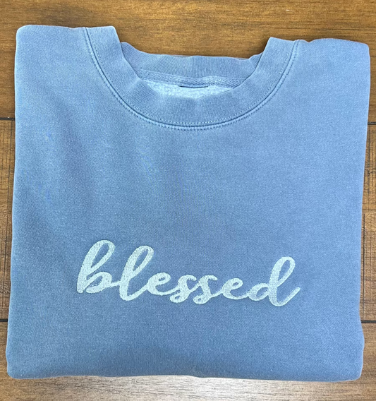 A folded light blue crewneck sweatshirt with the word 'blessed' embroidered in light blue script across the chest, laid flat on a brown wooden surface.