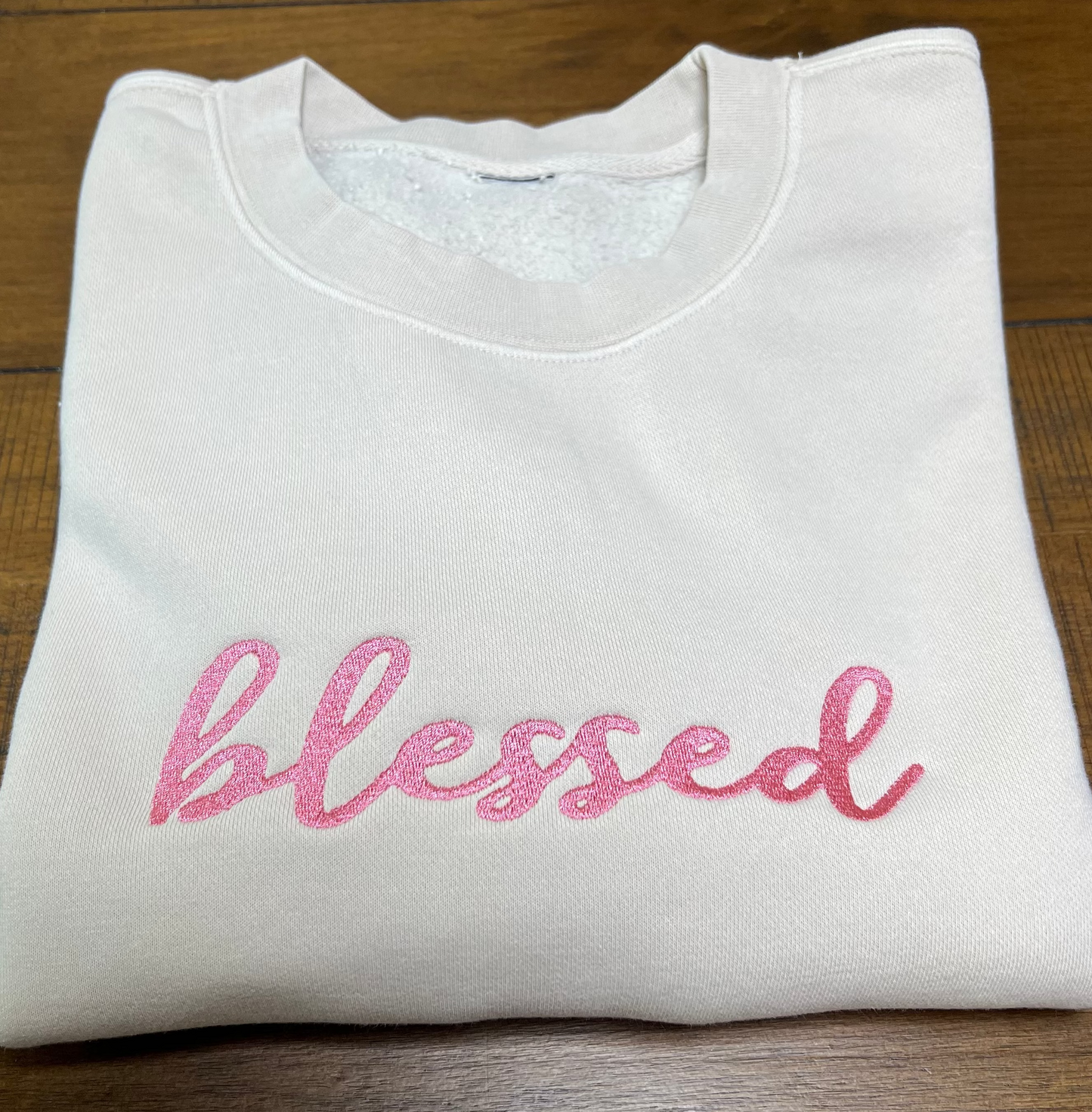 A folded cream crewneck sweatshirt with the word 'blessed' embroidered in pink script across the chest, laid flat on a wooden surface.