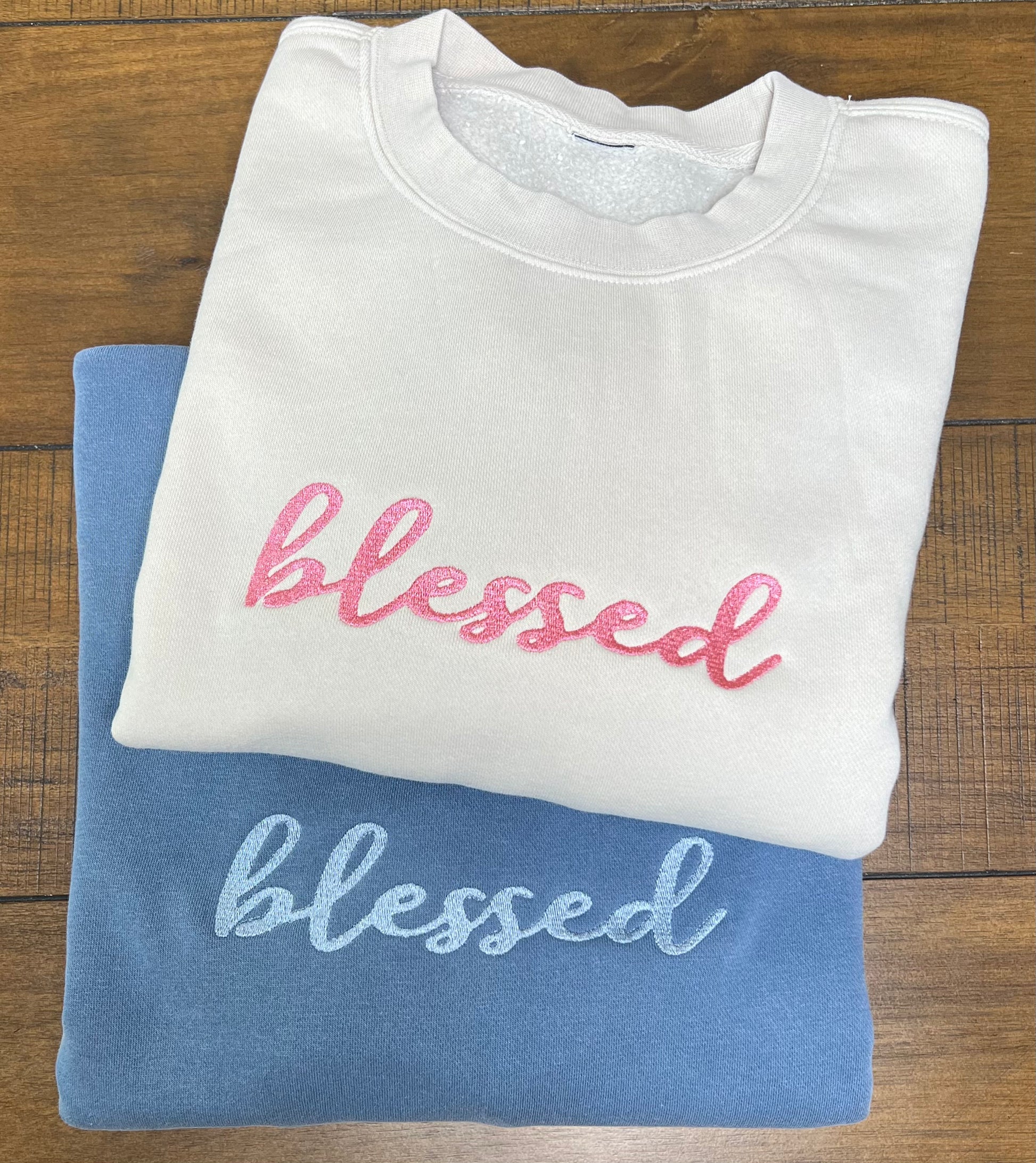 Two folded crewneck sweatshirts stacked on a wooden surface, one in white with pink embroidered 'blessed' and the other in blue with light blue embroidered 'blessed'.