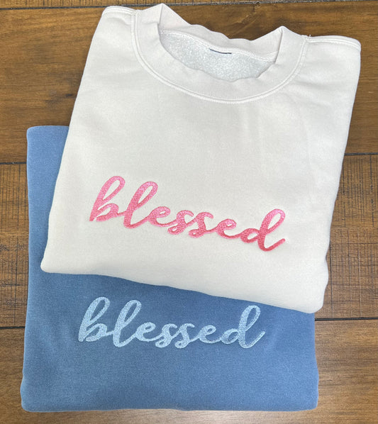 Two folded crewneck sweatshirts stacked on a wooden surface, one in white with pink embroidered 'blessed' and the other in blue with light blue embroidered 'blessed'.